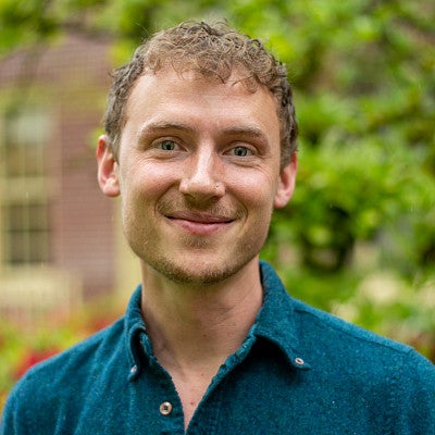 Photograph of Joe Wheeler, Second Year PhD Student in PPPM. Photo shows a masc-presenting individual in a teal collared shirt with short cropped hair and fair complexion, smiling into the camera. 